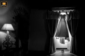Château des Girards, France, a bridal gown hangs dramatically in a brightly lit window, its edges glowing from the sun, while a small, glowing lamp offers a contrasting warm light across the frame.